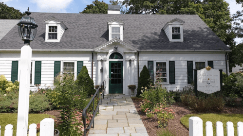 A charming white house with green shutters, a stone pathway, and a sign in the garden, surrounded by colorful flowers.