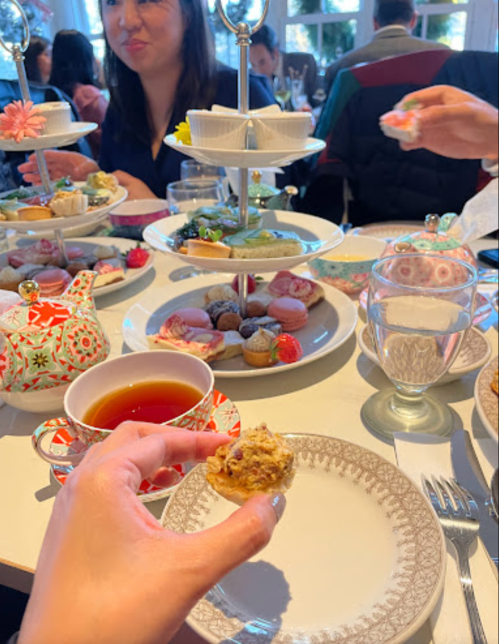 A hand holds a small pastry at a table set for afternoon tea, featuring tiered trays of desserts and tea cups.