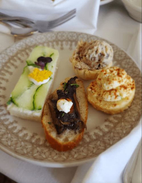 A plate with four small gourmet appetizers: cucumber sandwich, mushroom toast, seafood salad, and a creamy pastry.