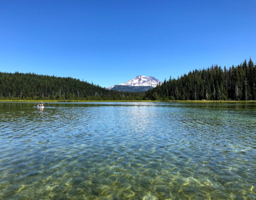 A clear lake reflects a blue sky and a snow-capped mountain, surrounded by lush green forests.