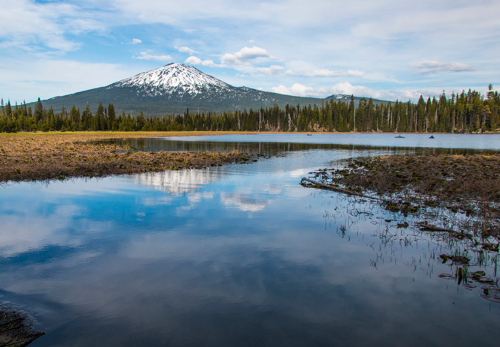A serene lake reflects a snow-capped mountain surrounded by lush forests under a blue sky with scattered clouds.