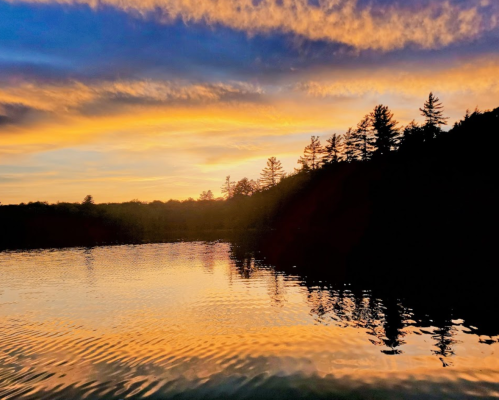 Sunset over a calm lake, with silhouettes of trees against a colorful sky of orange, pink, and blue hues.