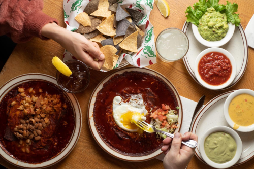 A table with two plates of enchiladas, chips, guacamole, salsa, and drinks, with hands reaching for food.
