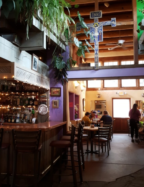 A cozy restaurant interior with a bar, plants, and patrons dining under wooden beams and a decorative cross.