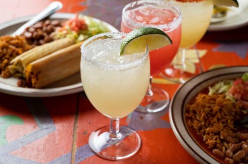 Three colorful cocktails with lime garnishes, alongside plates of tamales and rice on a vibrant table.