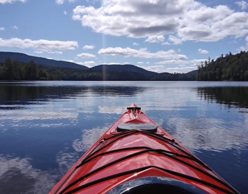 View from a red kayak on calm water, surrounded by mountains and a blue sky with fluffy clouds.