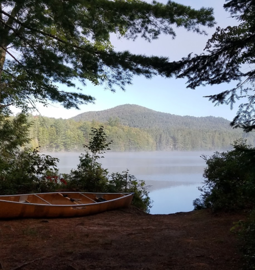 A serene lake view framed by trees, with a canoe on the shore and mist rising over the water and mountains in the background.