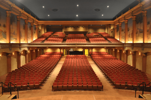 A spacious theater interior with rows of red seats and decorative walls, illuminated by soft lighting.