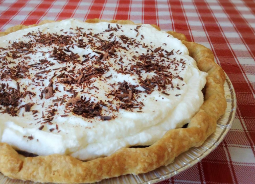 A creamy pie topped with whipped cream and chocolate shavings, sitting on a checkered red and white tablecloth.