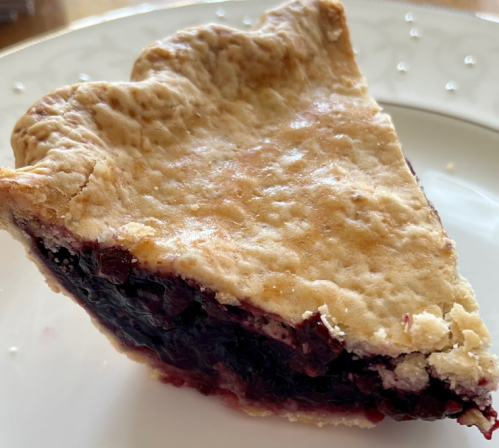 A slice of pie with a golden crust and dark berry filling on a decorative plate.