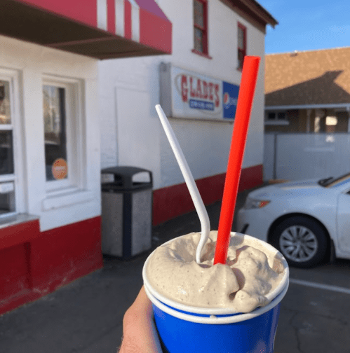 A hand holding a blue cup with a thick milkshake, featuring a red and a white straw, in front of a restaurant.