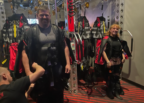 Two people in harnesses stand in a room with climbing gear, while a third person adjusts one of their harnesses.