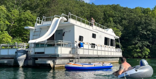 A houseboat with a slide on top, surrounded by trees, as a person in a small inflatable boat approaches.
