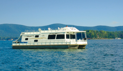 A houseboat floats on calm water with mountains in the background under a clear blue sky.