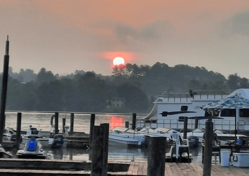 Sunset over a calm lake, with boats docked and silhouettes of trees in the background.