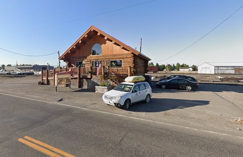 A log cabin-style building with a porch, parked cars in front, and a clear blue sky in the background.