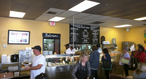A busy diner with customers at the counter, staff serving food, and a menu board on the wall.