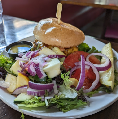 A burger on a plate with a side salad featuring lettuce, tomatoes, cucumbers, apples, and red onions.