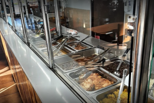 A food counter with various dishes in metal trays, each with serving utensils, in a casual dining setting.