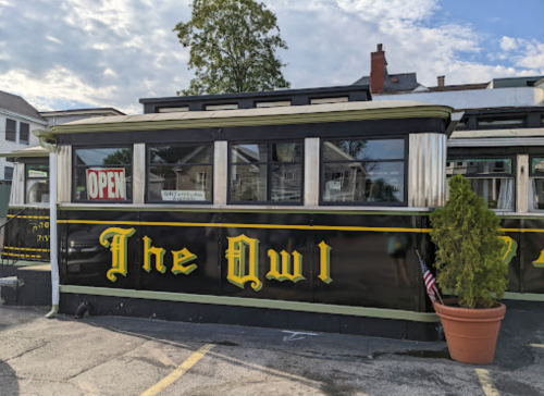 A vintage diner named "The Owl" with a black and yellow exterior, featuring an "Open" sign and a potted plant.