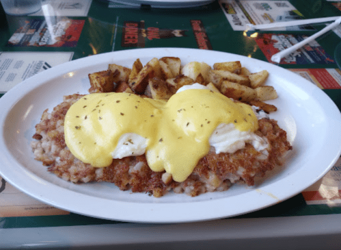 A plate of eggs Benedict on a crispy base, topped with hollandaise sauce, served with golden-brown potato wedges.