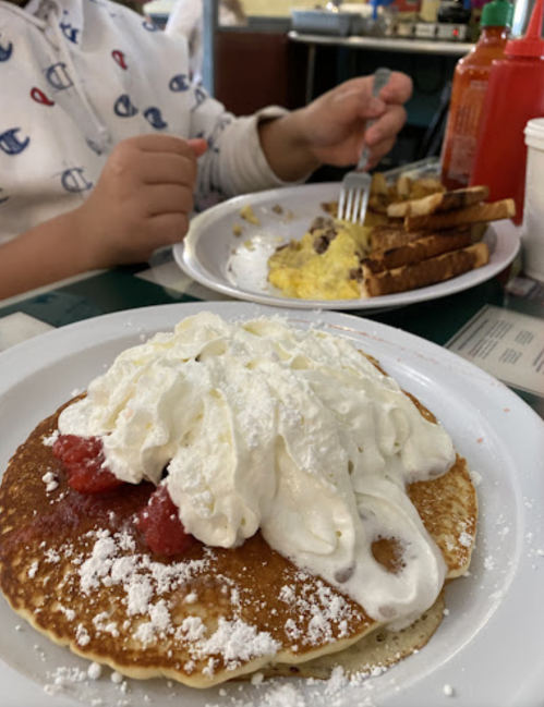 A plate of pancakes topped with whipped cream and strawberries, with a child’s hand holding a fork in the background.