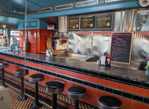 A retro diner interior featuring a counter with black stools, a menu board, and vintage decor.