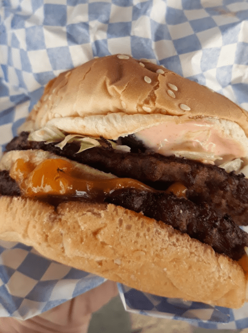 A close-up of a sesame seed bun burger with two patties, cheese, lettuce, and sauce, wrapped in blue checkered paper.