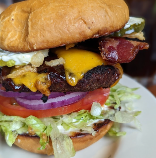 A close-up of a juicy burger with bacon, cheese, lettuce, tomato, pickles, and onions on a toasted bun.