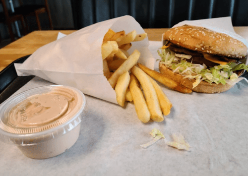 A sesame seed burger with lettuce and pickles, served with a side of fries and a small container of sauce.