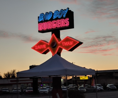Neon sign for "Bad Boy Burgers" with a drive-thru, set against a colorful sunset sky.