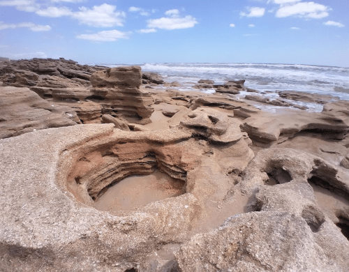 Rocky shoreline with unique formations and a sandy beach, under a blue sky with scattered clouds.
