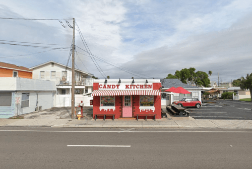 A colorful candy shop named "Candy Kitchen" with a red awning, surrounded by empty storefronts and a cloudy sky.