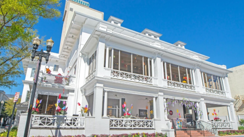 A white, historic building with large windows and colorful flowers, set against a clear blue sky.