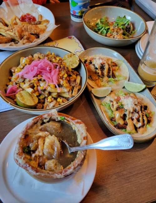 A table spread with various dishes: tacos, a bowl of soup, and a colorful corn salad with lime and pickled onions.