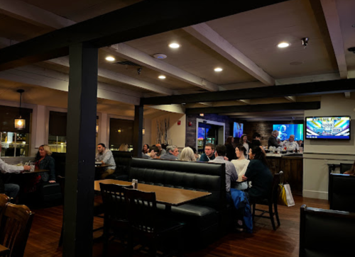A cozy restaurant interior with diners at tables, a bar area, and a TV showing a sports game in the background.