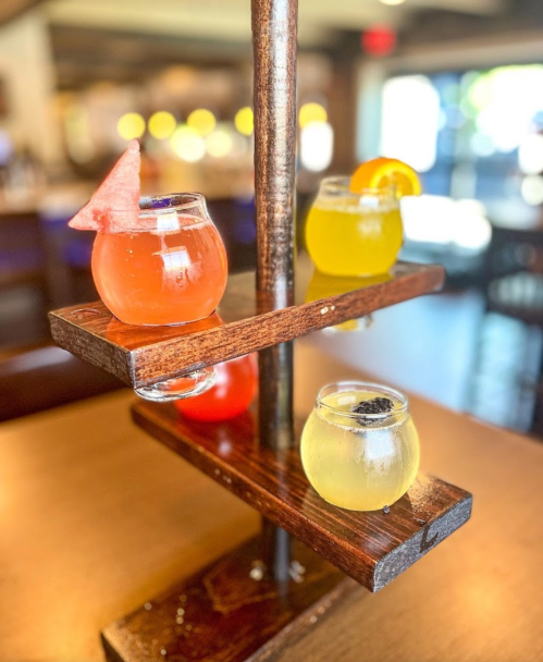 A wooden stand holds four colorful drinks in small glass bowls, featuring fruit garnishes and varying colors.
