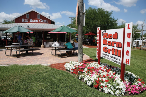 A colorful sign for Red Barn Grill outside a casual eatery with outdoor seating and flower beds.