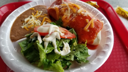 A plate with a red enchilada topped with cheese, a side of refried beans, and a fresh salad with tomatoes and dressing.