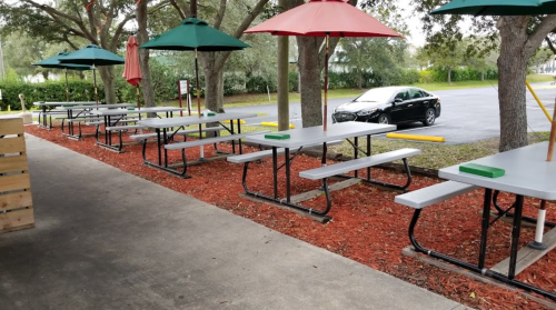 Outdoor seating area with tables and umbrellas, surrounded by trees and a parked car in the background.
