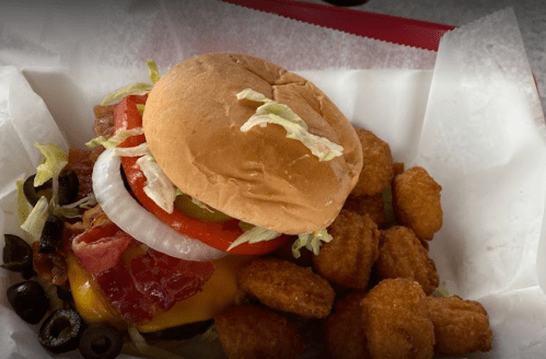A cheeseburger with bacon, lettuce, tomato, and onion, served with a side of crispy tater tots in a red basket.