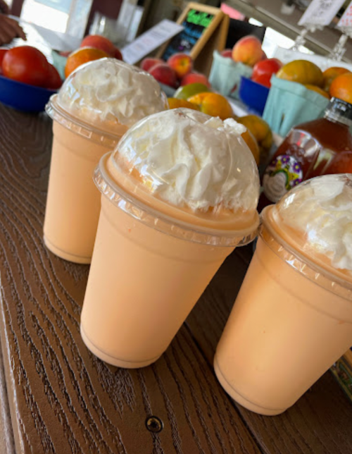 Three creamy orange drinks topped with whipped cream, placed on a wooden table with fresh fruit in the background.
