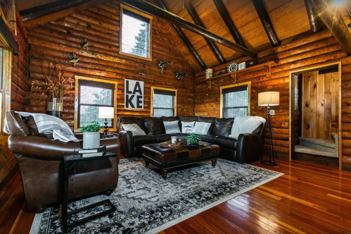 Cozy log cabin living room with leather sofas, a coffee table, and rustic decor, featuring large windows and wooden beams.