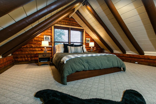 Cozy attic bedroom with a wooden ceiling, a large bed, and warm lighting, featuring rustic decor and carpeted flooring.