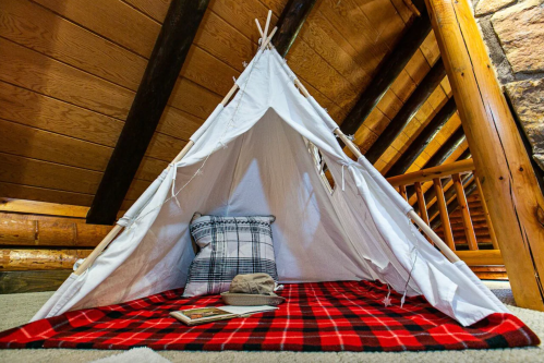 A cozy teepee tent with a plaid blanket and pillow, set in a rustic wooden cabin interior.
