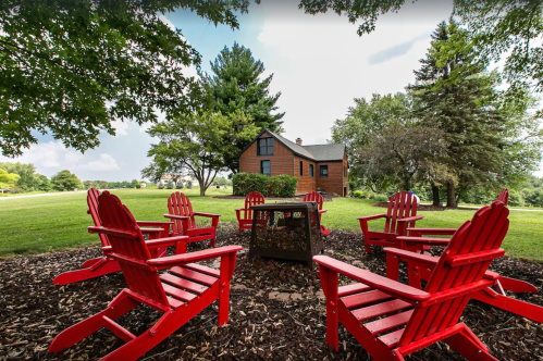 A cozy outdoor seating area with red chairs surrounding a fire pit, set against a rustic house and green landscape.