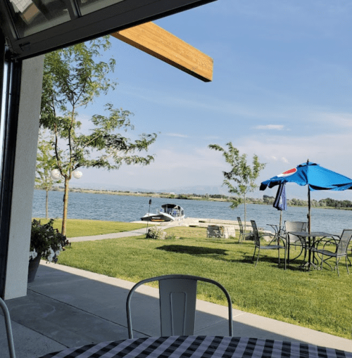 A serene lakeside view with green grass, trees, and a patio area featuring tables and an umbrella.