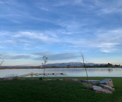 A serene lake view with a wooden dock, grassy foreground, and mountains under a blue sky with wispy clouds.