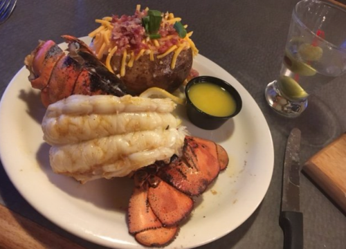 A plate featuring grilled lobster tails, a loaded baked potato, and a small glass with a lime garnish.