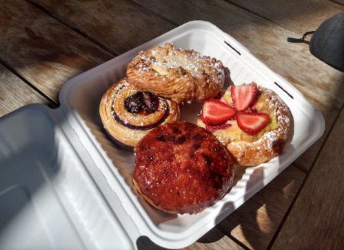A takeout container with four assorted pastries: a fruit tart, a chocolate croissant, and two sweet rolls.